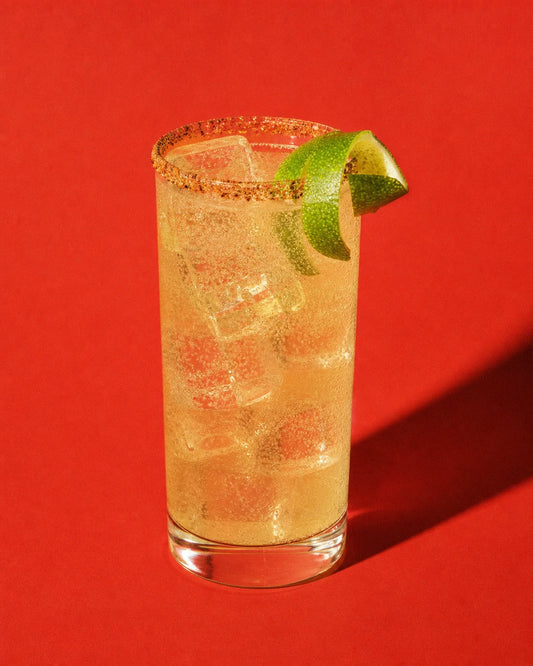 Acapulco Mule dorado pálido en vaso highball, con burbujas, twist de lima y Tajín sobre fondo liso.