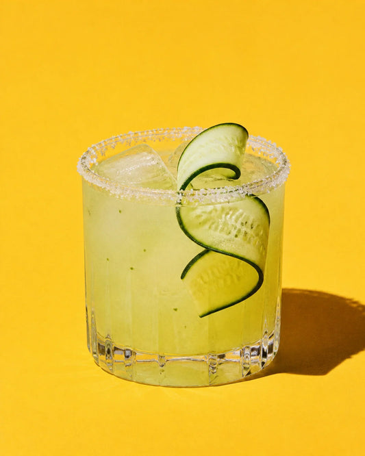Margarita Pepino verde pálido en vaso rocks, con una cinta larga de pepino sobre fondo liso.