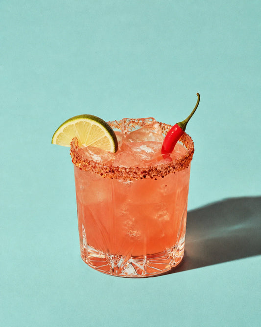 Margarita Picante de tono melocotón rojizo en vaso rocks, con borde de Tajín, lima y chile sobre fondo liso.