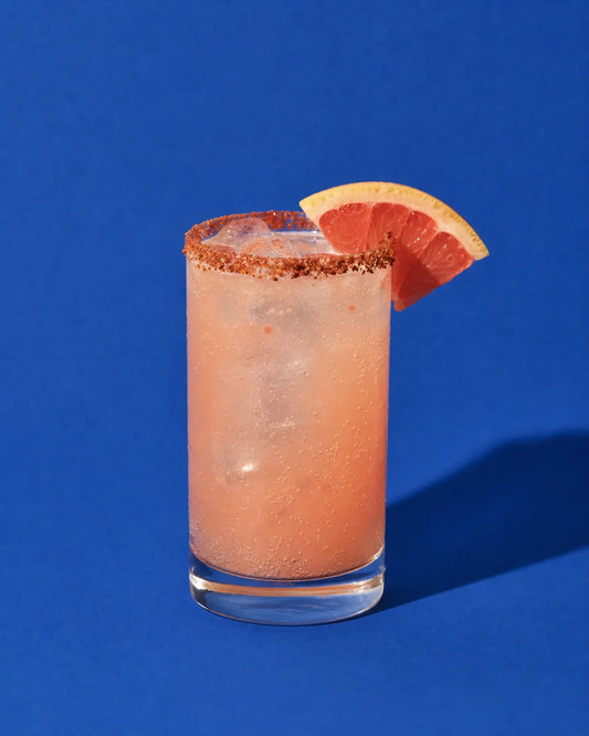 Paloma rosa suave en vaso highball, con borde de Tajín y cuña de pomelo sobre fondo liso.